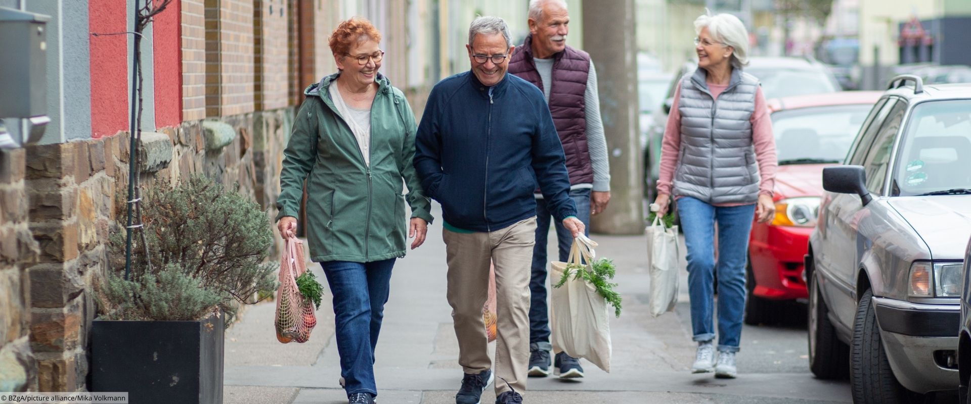 Vier ältere Erwachsener laufen gut gelaunt mit Einkaufstaschen einem Gehweg entlang.