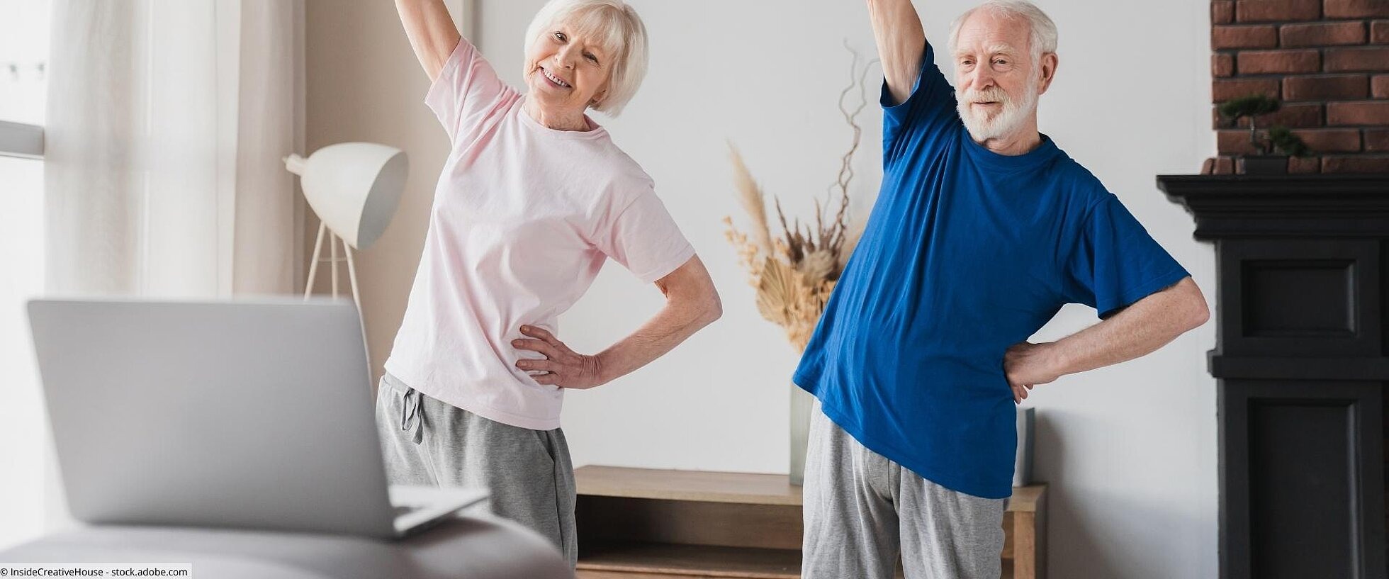Eine ältere Frau und ein älterer Mann bei Bewegungsübungen vor einem Laptop