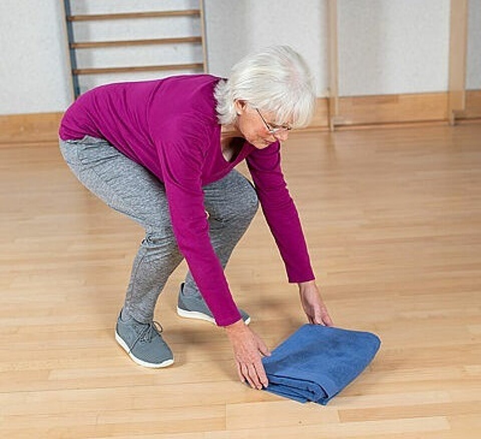 Kniebeugen-Übung: Frau geht in die Knie um ein Handtuch vom Boden zu heben.