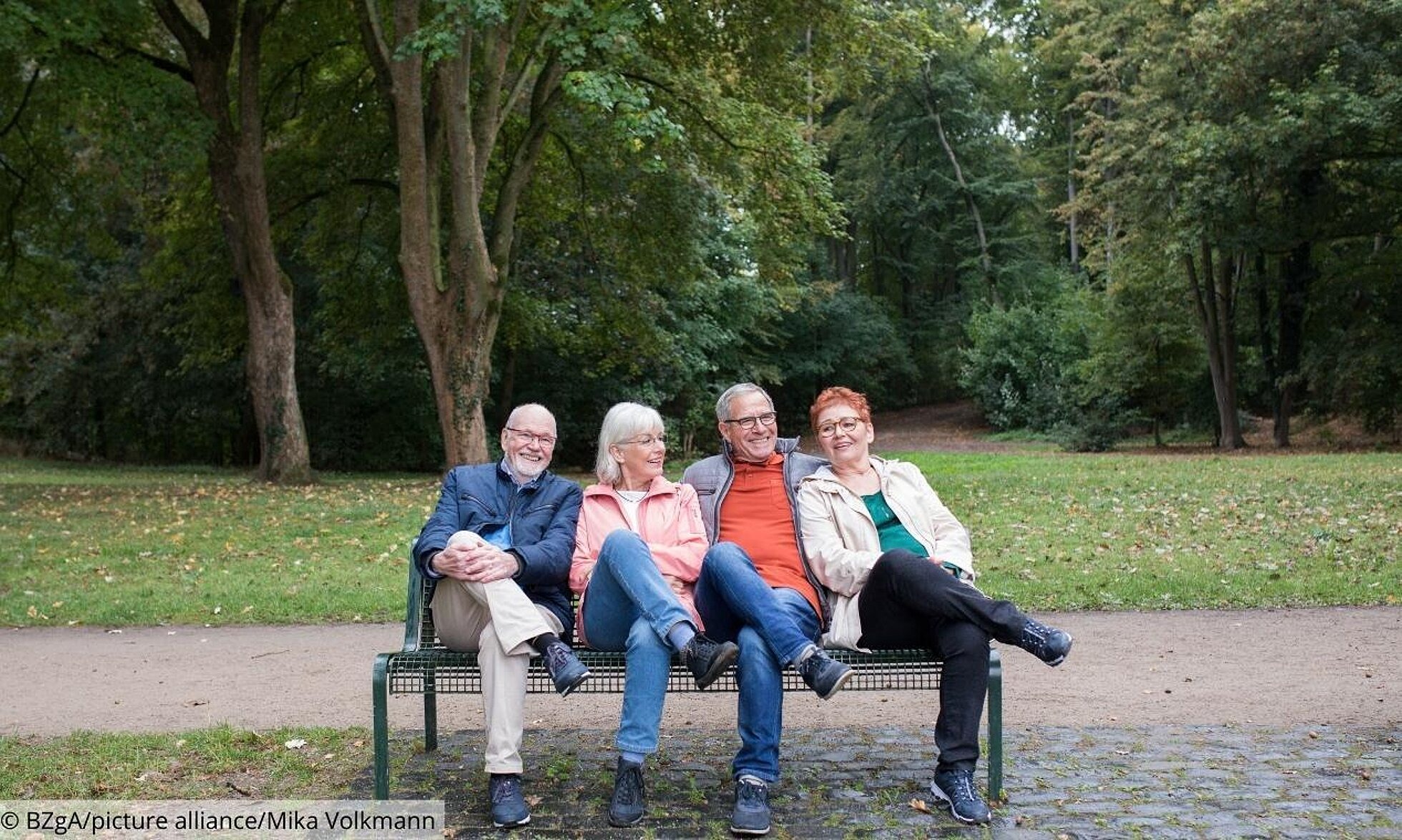 4 ältere Menschen sitzen auf einer Parkbank mit übereinandergeschlagenen Beinen.