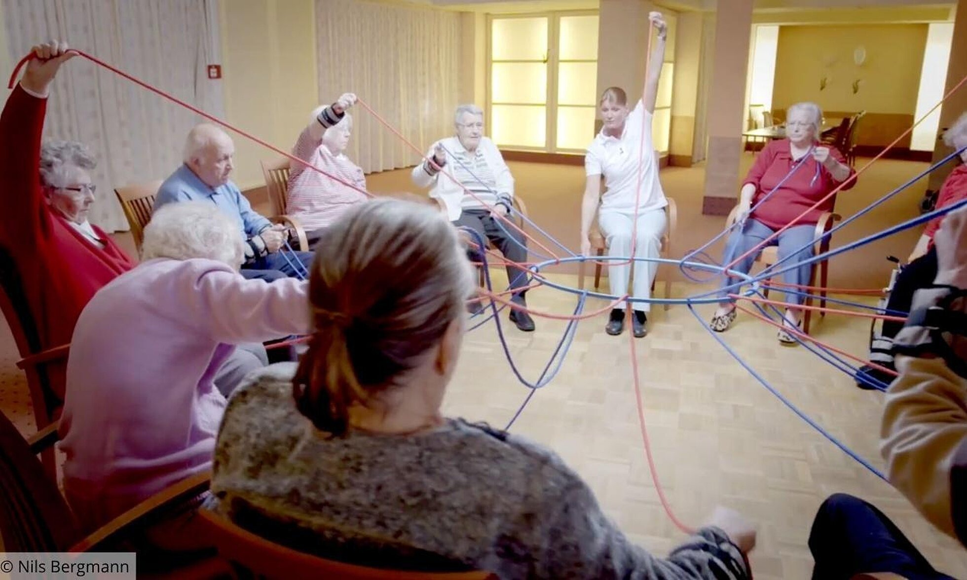 Gruppe von älteren Menschen hält mit Seilen einen Ring in der Mitte.