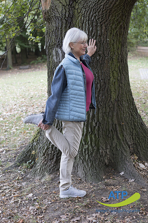 Ältere Frau dehnt Oberschenkel und stützt sich an einem Baum ab