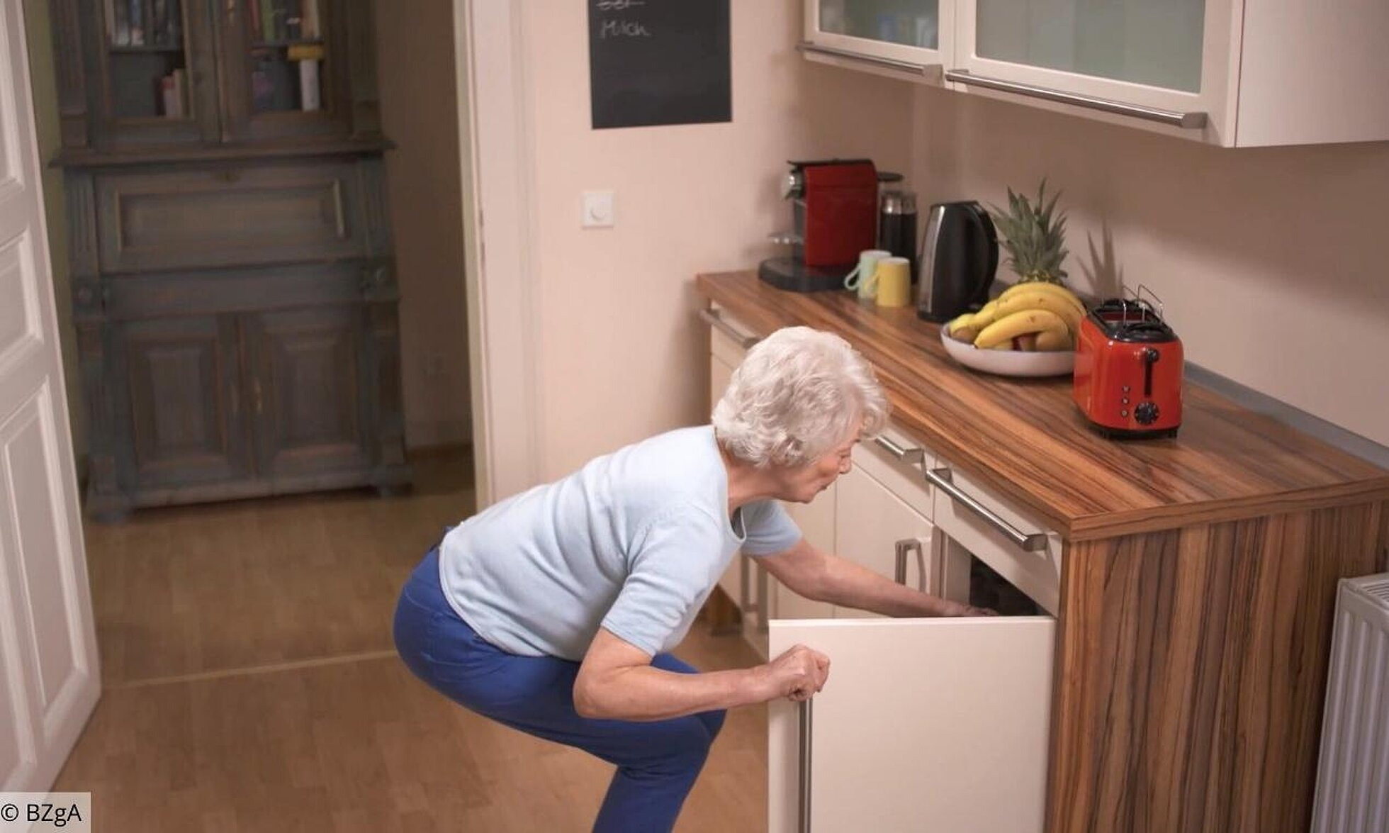 Ältere Frau geht in die Kniebeuge, um etwas aus dem unteren Küchenschrank zu holen