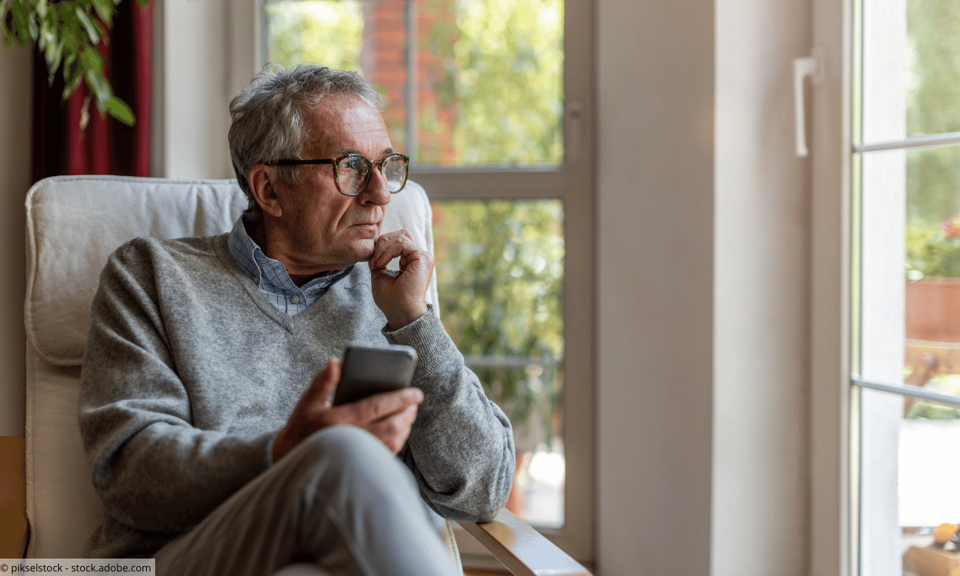 Älterer Mann mit Smartphone in der Hand schaut traurig aus dem Fenster